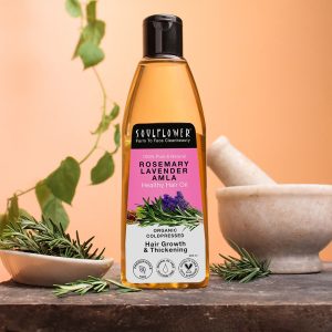 A bottle of "Soulflower Rosemary Lavender Hair Oil" placed on a wooden surface with rosemary sprigs and a mortar and pestle in the background. The label highlights that the oil is organic, cold-pressed, and promotes hair growth and thickening.