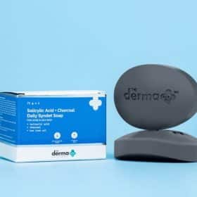 The Derma Co Salicylic Acid + Charcoal Daily Syndet Soap bar and packaging against a light blue background, emphasizing its use for acne-prone and oily skin.