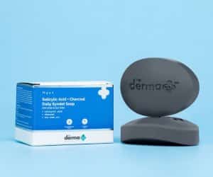 The Derma Co Salicylic Acid + Charcoal Daily Syndet Soap bar and packaging against a light blue background, emphasizing its use for acne-prone and oily skin.