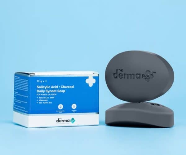 The Derma Co Salicylic Acid + Charcoal Daily Syndet Soap bar and packaging against a light blue background, emphasizing its use for acne-prone and oily skin.